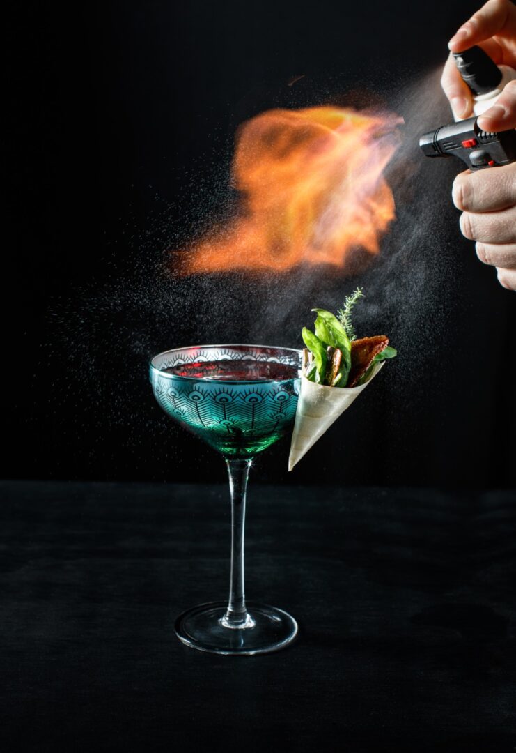 A person holding a cocktail in front of a flaming glass.